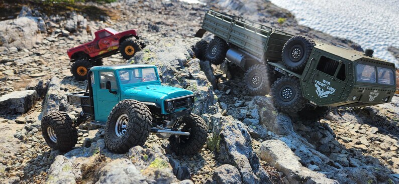 Pu&ograve; includere: Tre veicoli fuoristrada giocattolo su un terreno roccioso. Un camion blu turchese con pneumatici grandi, un camion rosso con un cassone nero e un camion militare verde con una piattaforma.