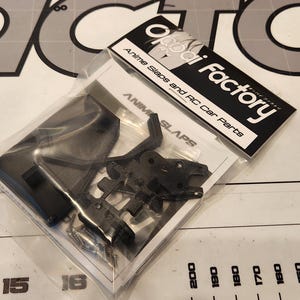 Puede incluir: Primer plano de una bolsa de pl&aacute;stico que contiene piezas de pl&aacute;stico negro para un anime slap y un coche RC. La bolsa est&aacute; etiquetada con "ai Factory" y "Anime Slaps and RC Car Parts".