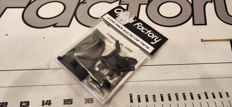 Puede incluir: Primer plano de una bolsa de pl&aacute;stico que contiene piezas de pl&aacute;stico negro para un anime slap y un coche RC. La bolsa est&aacute; etiquetada con "ai Factory" y "Anime Slaps and RC Car Parts".