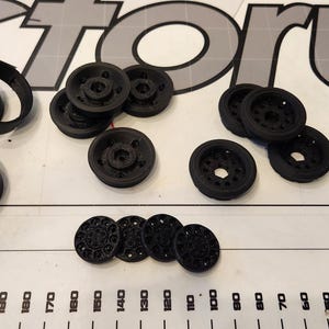 Pu&ograve; includere: Assortimento di ruote, pneumatici e viti in plastica nera. Le ruote hanno dimensioni e design diversi. Le viti sono piccole e argentate.