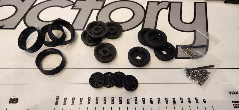 Pu&ograve; includere: Assortimento di ruote, pneumatici e viti in plastica nera. Le ruote hanno dimensioni e design diversi. Le viti sono piccole e argentate.