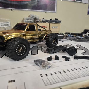 以下が含まれることがあります： 大きな黒いタイヤとロールバーを備えた、金色の玩具トラック。いくつかの黒いプラスチック部品と小さなハードウェアバッグが近くに配置されています。トラックは印刷されたマークのある表面にあります。