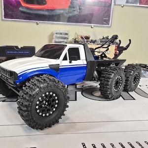 以下が含まれることがあります： 白と青のRCトラックモデル。 大きな黒いオフロードタイヤが付いています。 トラックは6輪構成で、牽引ヒッチが付いています。 モデルは、白いキャブ、青いアクセント、黒いシャーシで詳細に作られています。
