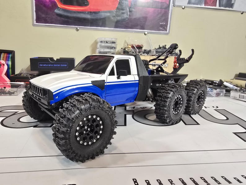 以下が含まれることがあります： 白と青のRCトラックモデル。 大きな黒いオフロードタイヤが付いています。 トラックは6輪構成で、牽引ヒッチが付いています。 モデルは、白いキャブ、青いアクセント、黒いシャーシで詳細に作られています。