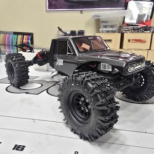 Puede incluir: Un camión teledirigido gris oscuro con neumáticos grandes y negros con tacos. El camión tiene una carrocería detallada, que incluye una cabina y una caja. La palabra "APEX" es visible en el lateral. El camión está sobre una superficie blanca con números y letras.