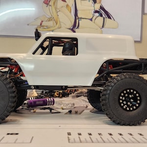 Puede incluir: Coche RC blanco con ruedas negras y una jaula antivuelco negra. El coche tiene una carrocer&iacute;a blanca y un interior negro. El coche est&aacute; sentado sobre una superficie blanca.