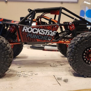 K&ouml;nnte beinhalten: Ein roter und schwarzer ferngesteuerter Rock-Crawler mit einem "Rockstar Punched"-Design an der Seite. Das Auto hat schwarze R&auml;der mit roten Akzenten und einen schwarzen &Uuml;berrollk&auml;fig.