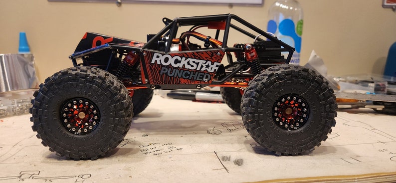 K&ouml;nnte beinhalten: Ein roter und schwarzer ferngesteuerter Rock-Crawler mit einem "Rockstar Punched"-Design an der Seite. Das Auto hat schwarze R&auml;der mit roten Akzenten und einen schwarzen &Uuml;berrollk&auml;fig.