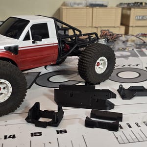 May include: A white and red toy truck with large, black tires and white rims. Several black plastic parts and a bag of screws are next to the truck. The truck has a roll cage and is on a white surface.