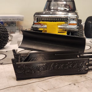 May include: A black plastic snow plow attachment for a toy truck. The plow is in two pieces and has the words "Custom Factory" printed on it.