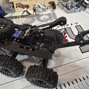 以下が含まれることがあります： 黒いオフロード車のシャーシと、大きく、ごつごつしたタイヤ。スペアタイヤが後部に搭載されています。車両は複雑なサスペンションシステムと赤いコンポーネントを備えています。車両は測定マークのある白い表面にあります。