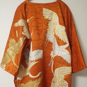 以下が含まれることがあります： オレンジ色の着物、鶴のデザイン。 白と金の鶴が羽を広げています。 背景には渦巻き模様があります。 着物はハンガーにかかっています。