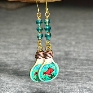 May include: Gold dangle earrings with a light blue glass bulb charm featuring a red fish and turquoise beads.