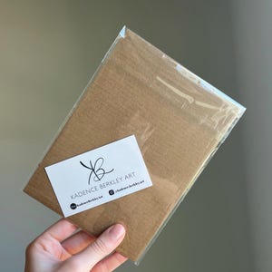 K&ouml;nnte beinhalten: Ein brauner Kunstdruck in einer transparenten Plastikh&uuml;lle. Ein wei&szlig;es Etikett mit dem Text "KADENCE BERKLEY ART" und Social-Media-Handles ist angebracht. Der Druck wird in einer Hand gehalten.