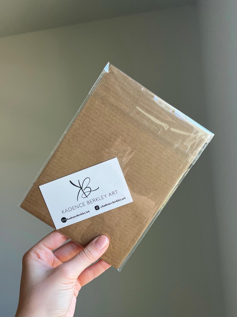 K&ouml;nnte beinhalten: Ein brauner Kunstdruck in einer transparenten Plastikh&uuml;lle. Ein wei&szlig;es Etikett mit dem Text "KADENCE BERKLEY ART" und Social-Media-Handles ist angebracht. Der Druck wird in einer Hand gehalten.