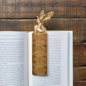 May include: A wooden bookmark featuring a fairy sitting on a book with the text "The Good Folk" on the cover. The bookmark is made of light brown wood and has a detailed design.