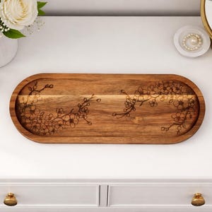 May include: A wooden, oval-shaped serving tray with a floral design etched into the surface. The tray is made of a light brown wood and is approximately 16 inches long. The floral design is a delicate, branch-like pattern.