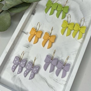 May include: A set of six pairs of handmade polymer clay earrings. The earrings are shaped like bows and come in four different colours: green, yellow, orange, and purple. The purple bows have white polka dots.