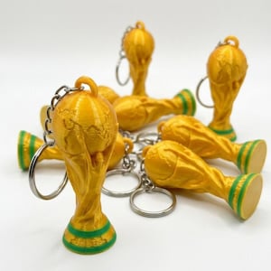 Könnte beinhalten: Eine Ansammlung goldfarbener Schlüsselanhänger in Form einer Trophäe mit grünen Akzenten. Die Schlüsselanhänger sind in Form der FIFA-Weltpokal-Trophäe gehalten. Auf weißem Hintergrund.