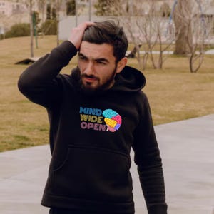 Op de afbeelding: Zwarte hoodie met de tekst "MIND WIDE OPEN" en een kleurrijke hersengrafiek. De sweatshirt heeft een voorzak en een capuchon met trekkoord. Het ontwerp gebruikt tinten blauw, geel, roze en groen.