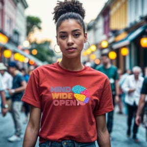 Puede incluir: Una camiseta de color óxido con las palabras "MIND WIDE OPEN" y un gráfico de cerebro colorido. La camiseta de manga corta es usada por una persona en una multitud.