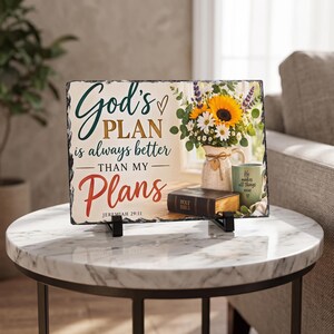 May include: A rectangular slate plaque featuring the text God's PLAN is always better THAN MY Plans JEREMIAH 29:11. The image shows a bouquet of sunflowers and lavender in a vase, a closed Holy Bible, and a green mug with the text He makes all things new.
