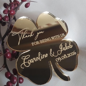 May include: A gold-colored, four-leaf clover-shaped wedding favor. The clover has the words "Thank you for being with us" and the names "Caroline & Jakob" along with the date "09.08.2026" engraved on it. The clover is next to a branch with red berries.