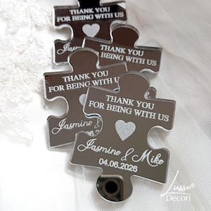 May include: Several silver puzzle piece favors with the text "THANK YOU FOR BEING WITH US" and a heart. One piece is engraved with "Jasmine & Mike 04.06.2026". The favors are from Lusso Decori.
