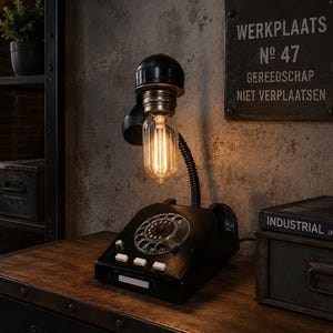 Op de afbeelding: Een bureaulamp in vintage-stijl met een zwarte telefoonvoet en een gloeilamp in plaats van de hoorn. De lamp heeft een retro-ontwerp, met een draaiknop en knoppen. De tekst op het bord luidt "WERKPLAATS No 47 GEREEDSCHAP NIET VERPLAATSEN."