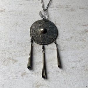 May include: A silver necklace with a round pendant featuring an intricate floral design. The pendant has three dangling silver charms. A clear crystal bead hangs above the pendant.