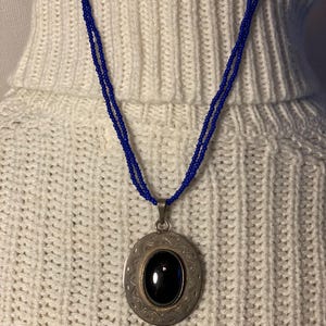 Può includere: Una collana con un ciondolo in argento con una pietra ovale nera incastonata in una cornice argentata dettagliata. La collana è realizzata con perline blu.