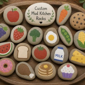 May include: Hand-painted rocks with food illustrations, including an apple, carrot, cookie, and watermelon. The rocks are arranged in a wooden bowl. One rock has the text "Custom Mud Kitchen Rocks".