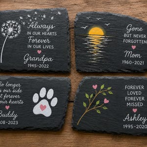 May include: Four black slate memorial stones with white and pink painted designs and text. One stone says "Always in our hearts, Forever in our lives, Grandpa 1945-2022". Another says "Gone but never forgotten, Mom 1960-2021".