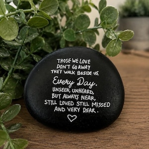 May include: A smooth, round black stone featuring white hand-painted text that reads: THOSE WE LOVE DON'T GO AWAY THEY WALK BESIDE US Every Day. UNSEEN, UNHEARD, BUT ALWAYS NEAR, STILL LOVED STILL MISSED AND VERY DEAR. A small white heart is below.