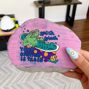 May include: A hand-painted rock with a frog in a bathtub design. The frog is green, the tub is white, and the rock has a pink background. Text on the rock reads "splish splash" and "your opinion is trash".
