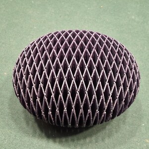 May include: A dark purple, spherical object with a lattice design. The object is made up of intersecting diamond shapes, creating a textured surface. The item is sitting on a green surface.