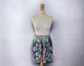 Pode incluir: Uma saia curta com estampa floral com tulipas rosa, amarelas e azuis sobre um fundo azul escuro. A saia tem uma cintura com cordão rosa.