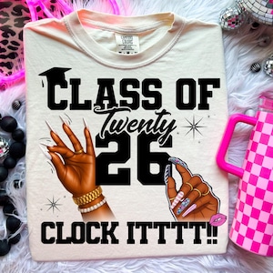 May include: Cream-colored t-shirt with the text "CLASS OF Twenty 26 CLOCK ITTTT!!" in black. The design includes illustrations of hands with long, decorated nails, a graduation cap, and a star. A pink checkered tumbler is visible.