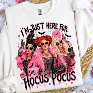 May include: A white long-sleeve sweatshirt featuring a graphic print of three women with pink feather boas and sunglasses. The text reads I'M JUST HERE FOR THE HOCUS POCUS. The design includes bats, stars, a skull, and a castle silhouette in pink and black.