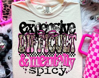 May include: Cream-colored t-shirt with the text "Expensive, Difficult & Mentally Spicy" in black, pink, and leopard print. A pink checkered tumbler with a handle is on the right. The shirt is on a white fluffy surface.
