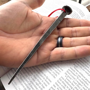 May include: A hand-forged iron nail with a square, tapered shaft and a flat, rounded head. A small hole is drilled through the head, threaded with a thin red ribbon. The metal has a dark, textured, gunmetal gray finish with visible hammer marks.