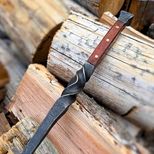 May include: A hand-forged fire poker with a twisted steel shaft and a dark wood handle. The handle is secured with brass rivets. The poker is designed for use with a fireplace or campfire.