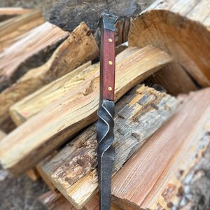May include: A handcrafted fire poker with a twisted steel shaft and a wooden handle. The handle is a rich, dark red with three brass rivets. The top of the poker is a square shape. The overall design is rustic and functional.