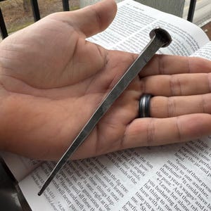 May include: A hand-forged iron nail with a dark, textured finish and a square-shaped shaft that tapers to a sharp point. The nail features a flat, slightly irregular head and a metallic gray surface, showcasing visible hammer marks along its length.