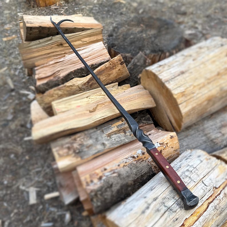 May include: A handcrafted, black metal fireplace poker with a curved hook at the end. The handle is made of dark wood and is secured with brass rivets. The poker is resting on a pile of firewood.