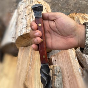 May include: A hand-forged metal and wood tool with a dark, twisted metal blade and a dark wood handle. The handle is secured with brass rivets. The tool has a square, metal head.