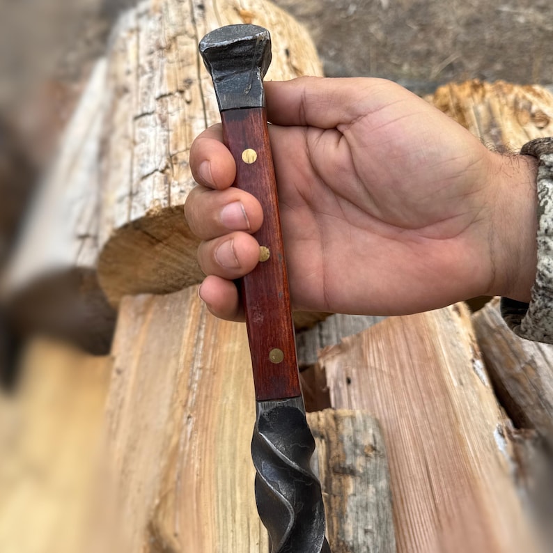 May include: A hand-forged metal and wood tool with a dark, twisted metal blade and a dark wood handle. The handle is secured with brass rivets. The tool has a square, metal head.