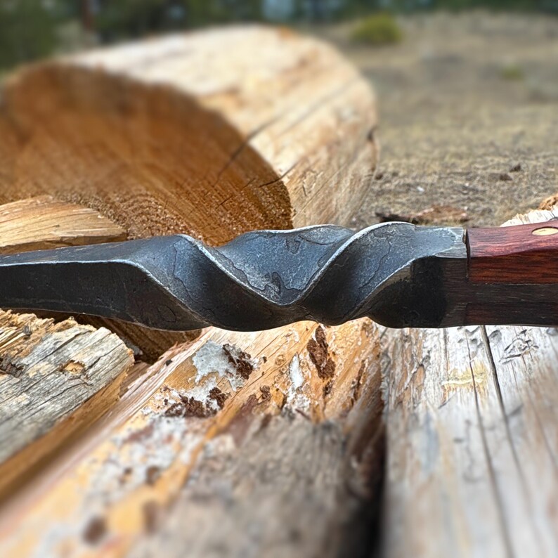 May include: Close-up of a hand-forged metal spearhead with a twisted design. The spearhead has a dark, textured finish and is attached to a wooden handle with a rich, reddish-brown color. The spearhead is resting on a wooden surface.