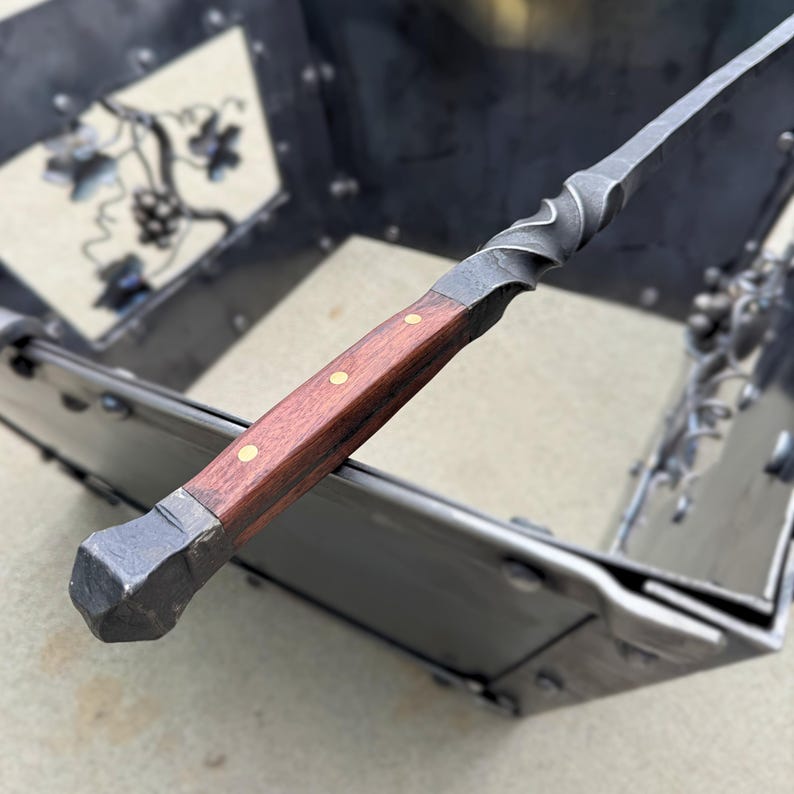 May include: Close-up of a hand-forged metal knife with a dark wood handle. The handle is secured with brass rivets. The blade has a twisted design near the handle. The knife is part of a metal decorative piece.