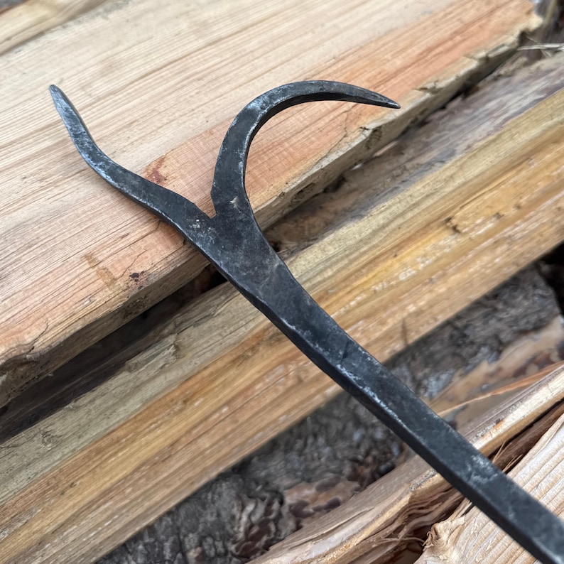 May include: A black, forged metal fireplace poker with a curved hook and a forked end. The tool is set against a backdrop of stacked firewood, showcasing its rustic design and intended use for managing a fire.